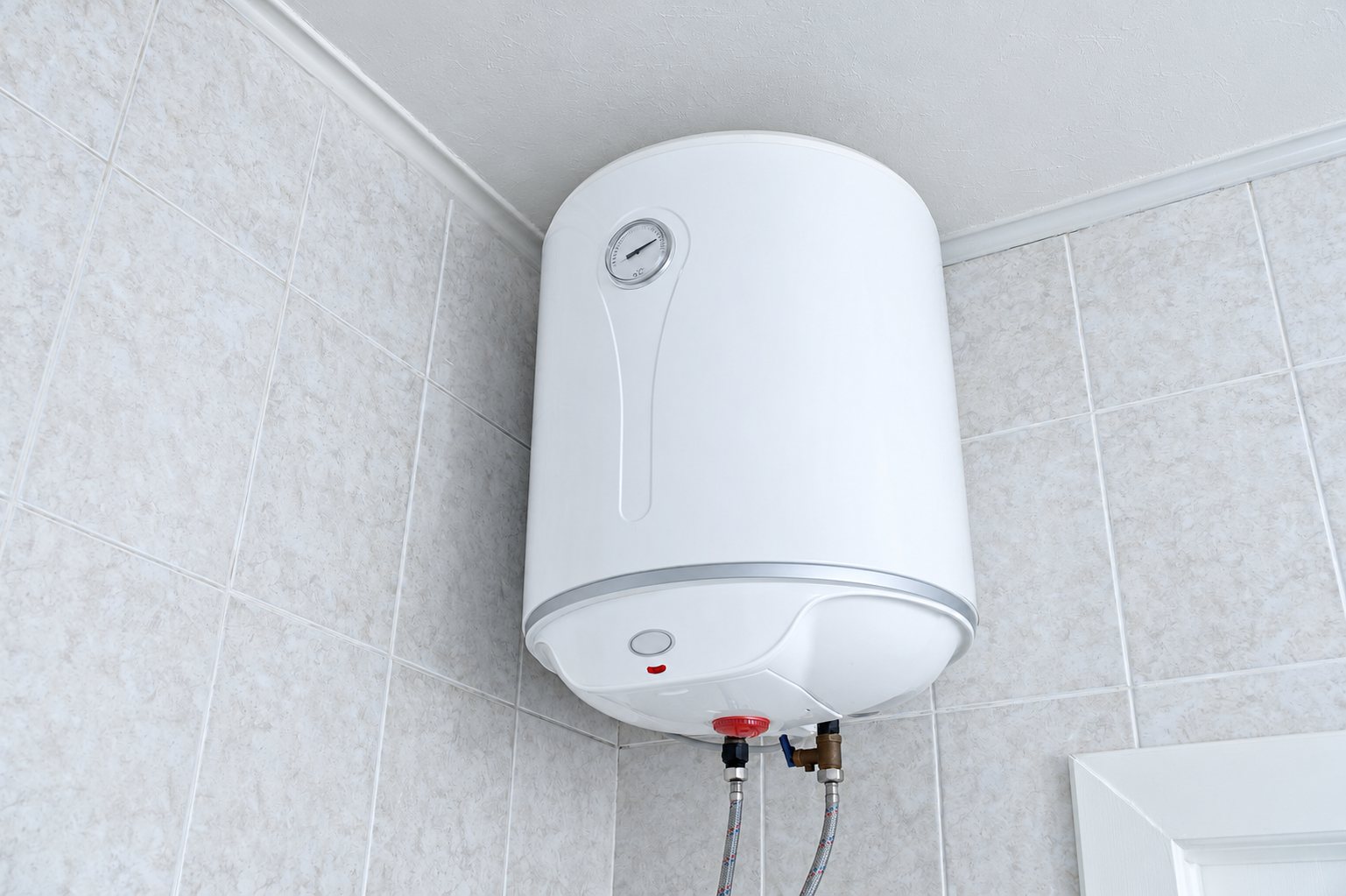 Un chauffe-eau électrique blanc installé dans l'angle d'une salle de bain carrelée, avec ses flexibles de raccordement visibles en dessous.