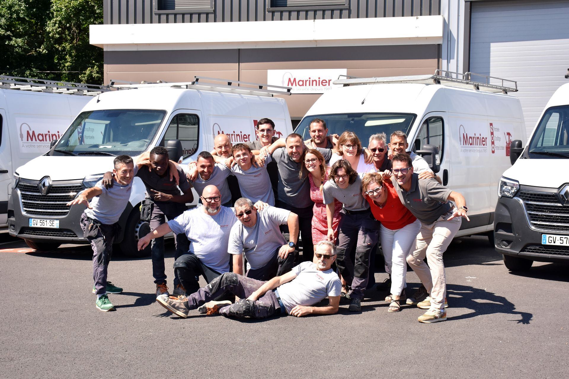 Une quinzaine de collègues posent en groupe, de façon joyeuse, sur un parking devant des camionnettes blanches portant le nom Marinier.