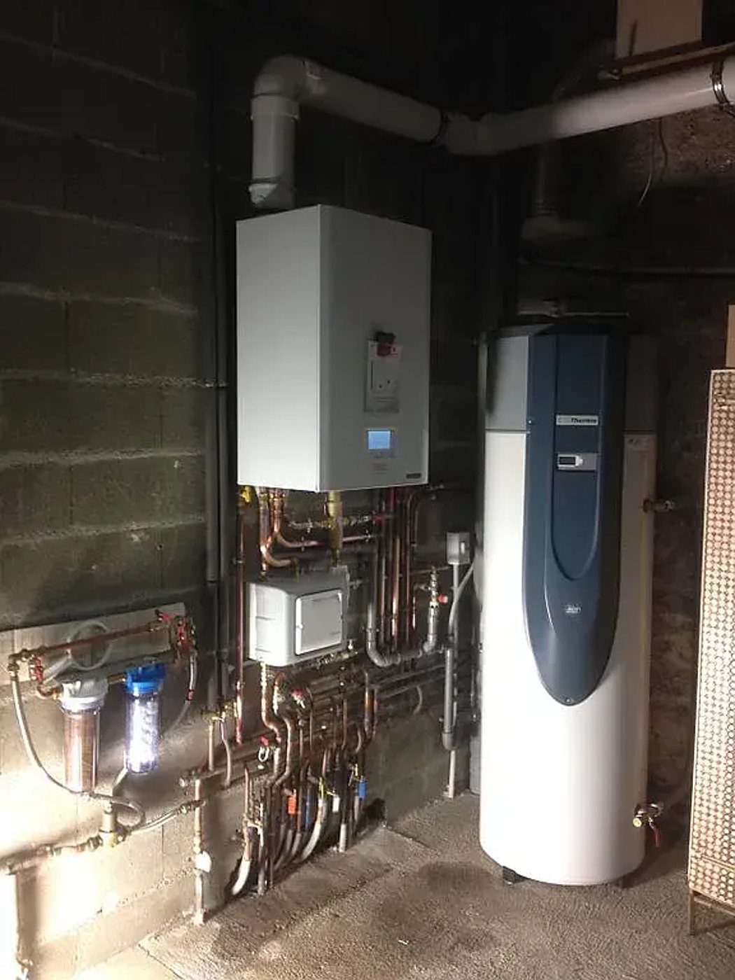 Dans un local technique aux murs en parpaing, une chaudière murale blanche, un réseau de tuyaux en cuivre, des filtres à eau et un grand ballon d'eau chaude Thermor bleu et blanc sont installés.