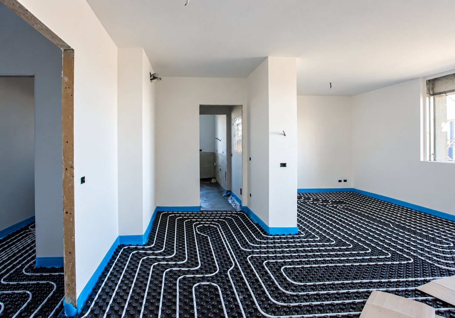 Pièces aux murs blancs en chantier dont le sol est couvert de plaques à plots noirs et de tuyaux de chauffage au sol blancs, avec une bande périphérique bleue.