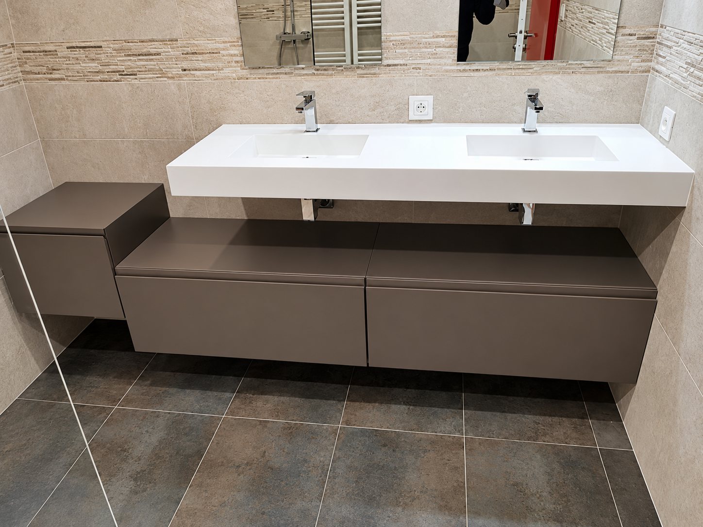 Salle de bain rénovée avec une double vasque blanche posée sur un meuble suspendu gris taupe à tiroirs, devant un mur en carrelage beige et un sol en carreaux gris foncé.