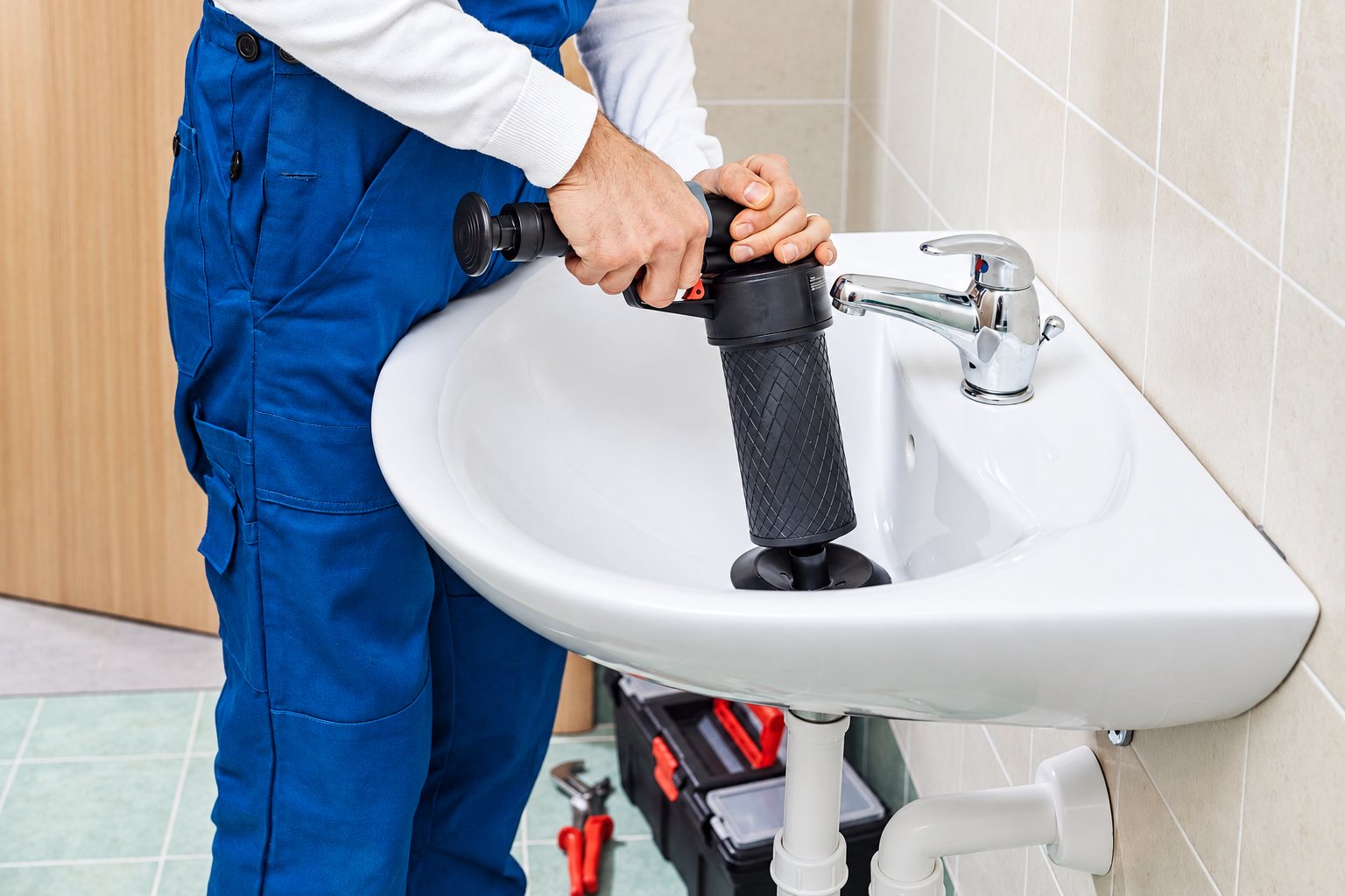 Un plombier en salopette bleue utilise une pompe à pression pour déboucher un lavabo blanc dans une salle de bains, avec sa caisse à outils au sol.