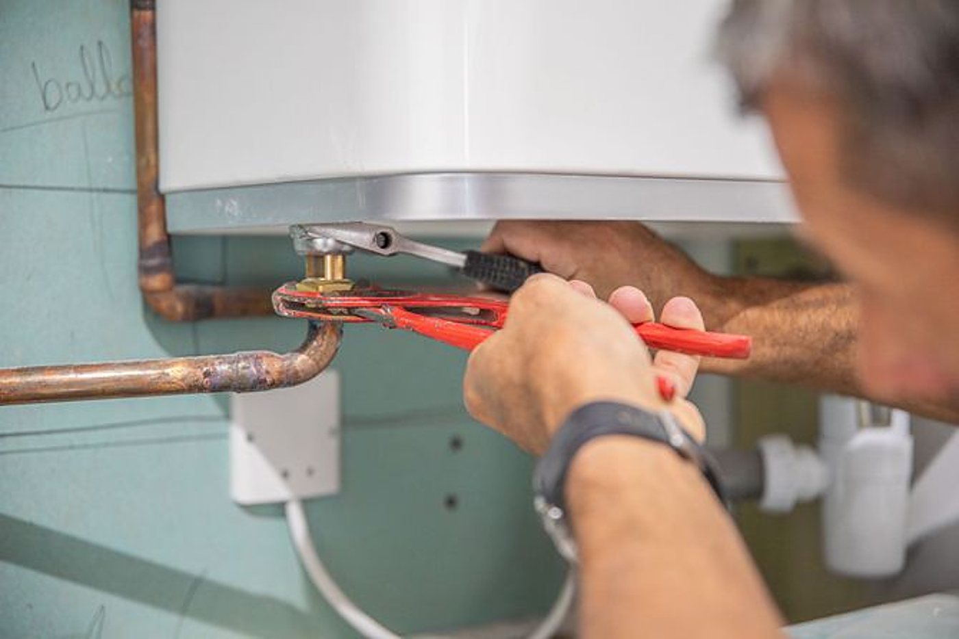 Un plombier utilise une pince multiprise rouge pour serrer un raccord en laiton sous un chauffe-eau mural, près de tuyaux en cuivre.