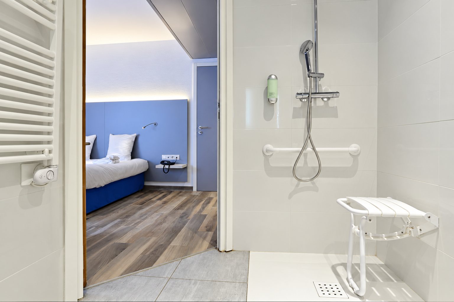 Salle de bain adaptée aux personnes à mobilité réduite avec douche de plain-pied, siège rabattable et barre d'appui, ouverte sur une chambre aux tons bleus.