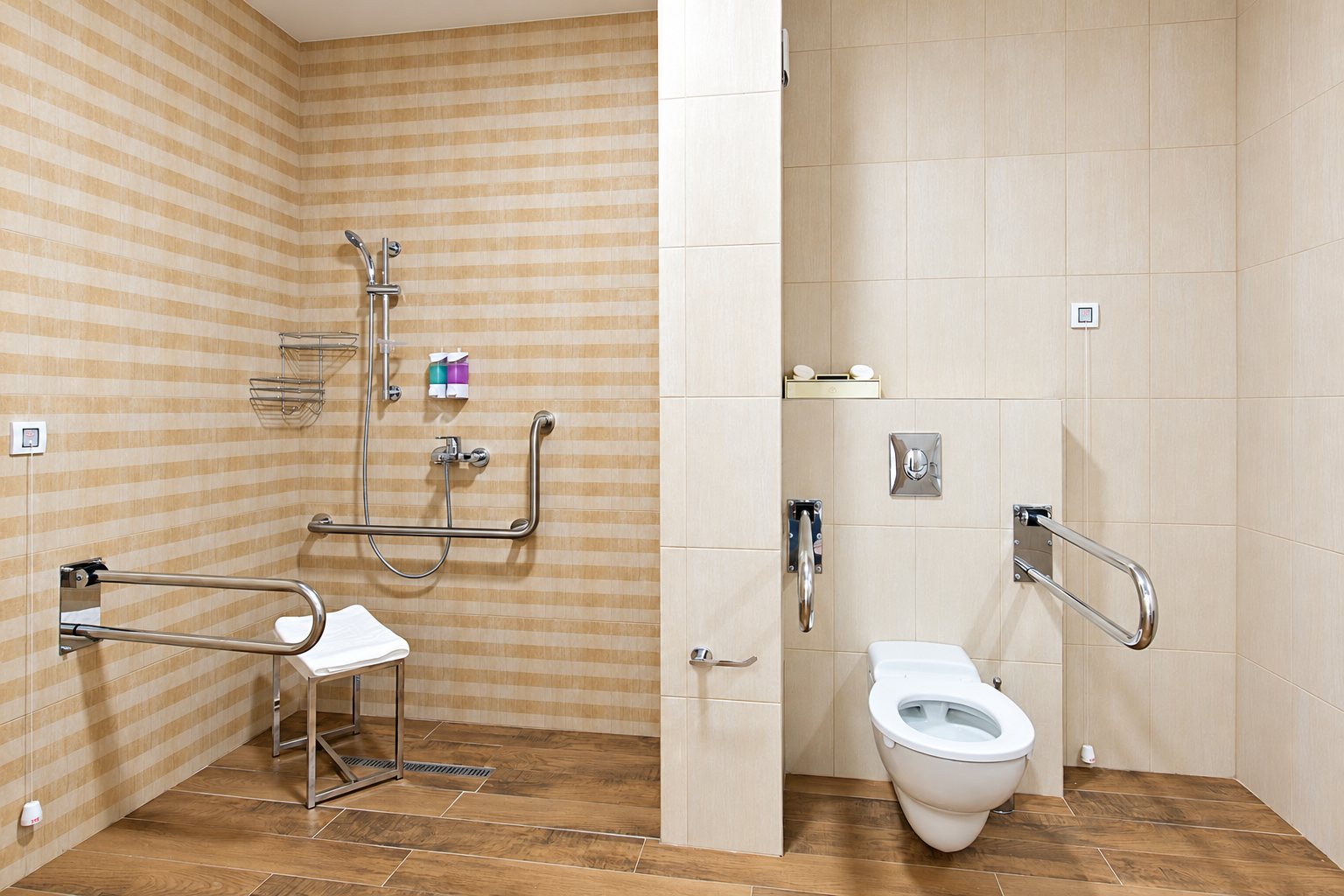 Salle de bain accessible aux personnes à mobilité réduite avec douche à l'italienne, siège, barres d'appui et WC suspendu.