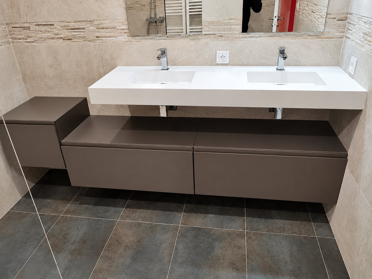 Salle de bain rénovée avec un plan double vasque blanc, des robinets chromés et un meuble suspendu taupe sur un carrelage sombre.